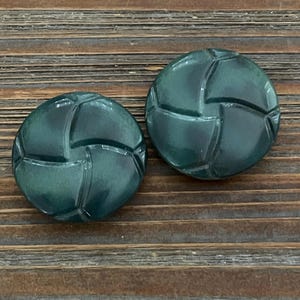 May include: Two dark teal, round buttons with a geometric design. The buttons have a glossy finish and are set against a wooden background. These buttons could be used for sewing or crafts.