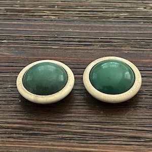 May include: Two round earrings with a dark green, oval-shaped center and a light beige border. The earrings are set against a dark brown wooden surface.
