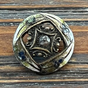 May include: A round, antique button with an ornate design. The button features a central, faceted gem surrounded by a square frame and intricate metalwork. The outer edge has a floral pattern with green and blue accents.