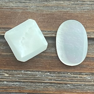May include: Two pale green gemstone cabochons. One is a square shape with beveled edges, and the other is an oval shape. The gemstones are set against a wooden background with horizontal planks.