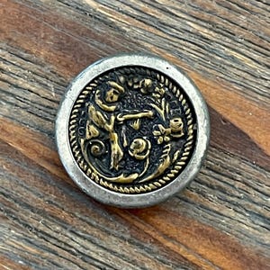 May include: A silver and gold button with a raised design of a person holding a flower. The button has a rope-like border.