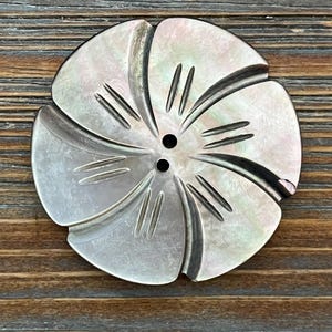 May include: A large, iridescent mother-of-pearl button in the shape of a flower. The button has six petals with etched lines and two small holes in the center. The button is set against a wooden background.