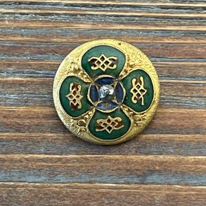 May include: A gold-toned button with a green, blue, and red enamel design. The button has a raised center with a small, clear stone.