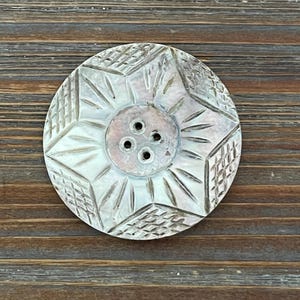May include: A circular, iridescent button featuring a central four-hole design surrounded by a star-shaped pattern. The surface displays alternating sections of etched, radiating lines and a cross-hatched grid texture across its pearlescent finish.