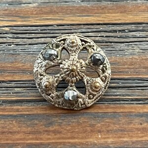 May include: A silver and gold decorative button with a floral design and four black rhinestones.