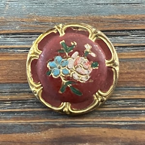 Puede incluir: Un botón redondo rojo con un borde dorado y un diseño floral. Las flores son rosas, azules y verdes.