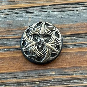 May include: A silver button with a decorative design featuring a star in the center and a woven pattern around it. The button has a black background and a small clear rhinestone in the center of the star.