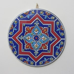 May include: A round ceramic wall hanging with a blue, red and white geometric design. The design features a large eight-pointed star with intricate floral patterns.