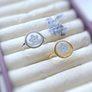 May include: Two rings with forget-me-not flowers encased in clear resin. One ring is silver and the other is gold.