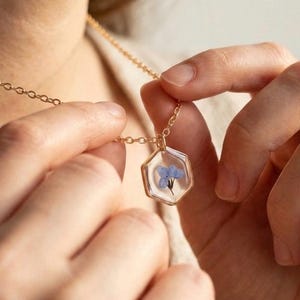 May include: A gold-colored necklace with a hexagonal pendant. The pendant encases a small, delicate blue flower in clear resin. The necklace is being held by a person.