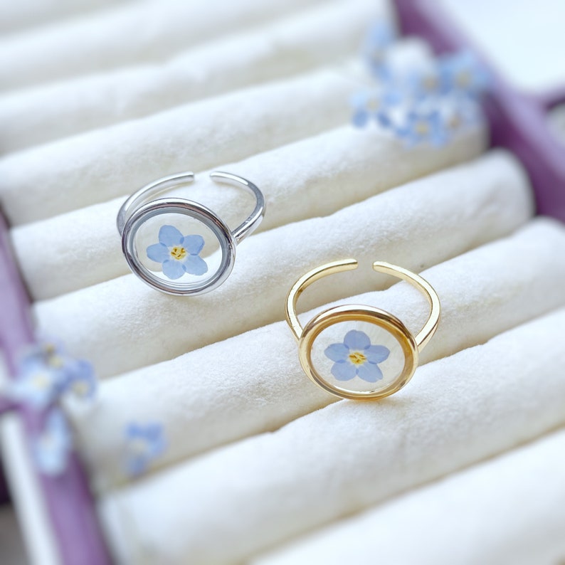 May include: Two rings with a forget-me-not flower encased in resin. The ring on the left is silver and the ring on the right is gold.