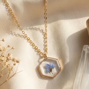 May include: A gold-colored necklace with a hexagonal pendant. The pendant is clear, encasing a small blue flower. The chain is delicate, and the necklace rests on a cream-colored, satin-like fabric. The overall aesthetic is delicate and floral.