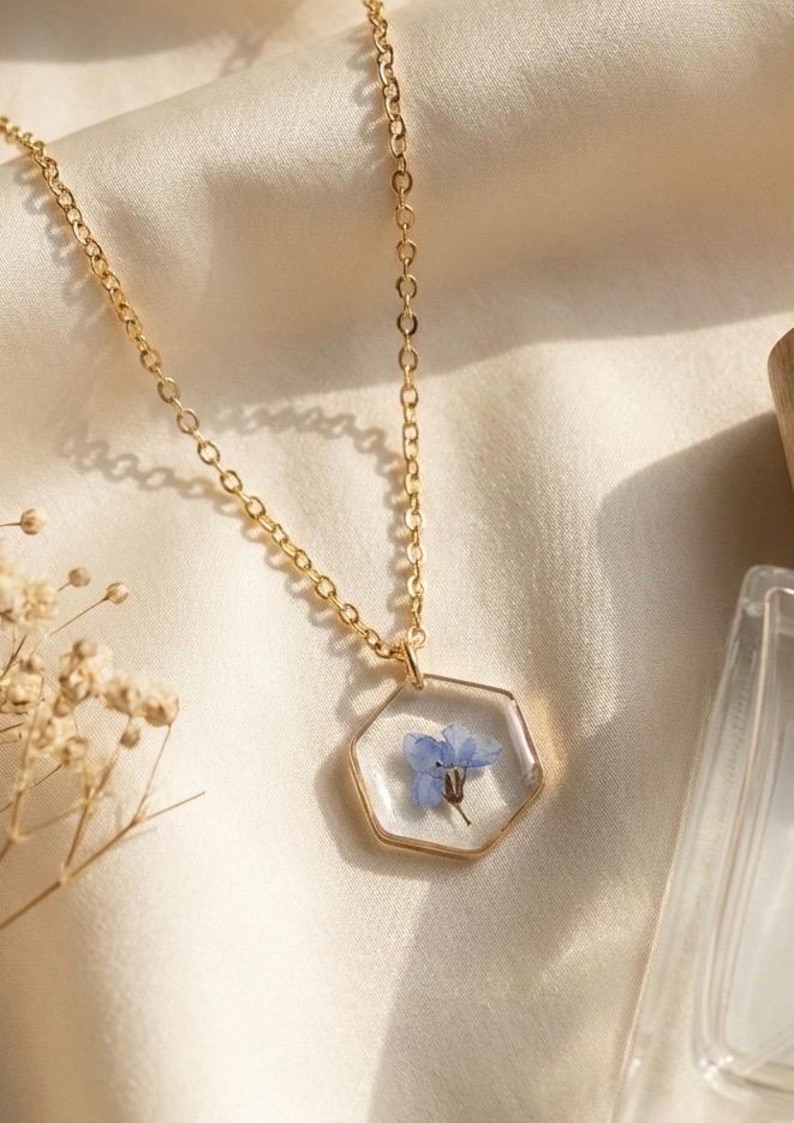 May include: A gold-colored necklace with a hexagonal pendant. The pendant is clear, encasing a small blue flower. The chain is delicate, and the necklace rests on a cream-colored, satin-like fabric. The overall aesthetic is delicate and floral.