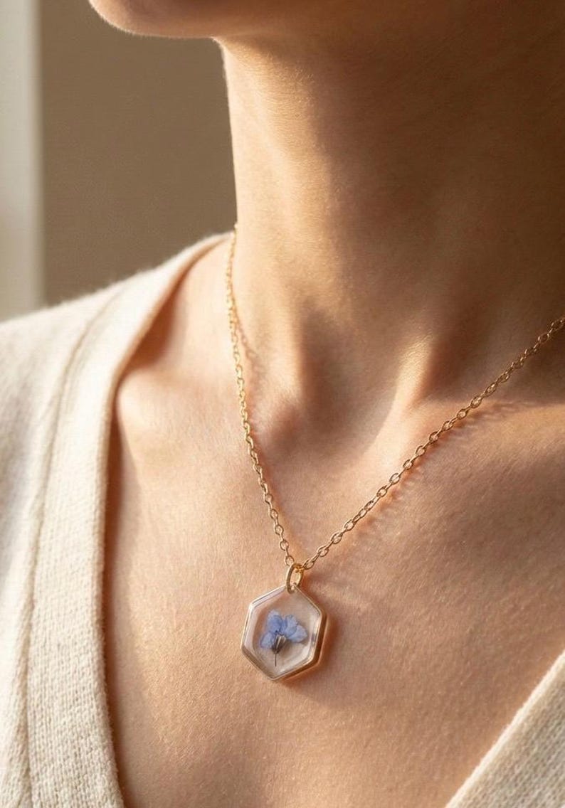May include: A gold-colored necklace with a hexagonal pendant. The pendant encases a small, delicate blue flower in clear resin. The necklace chain is fine and links together. The background is a soft, neutral color.