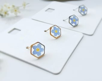 May include: A pair of stud earrings featuring a hexagonal design with a light blue flower and yellow center encased in a clear resin. The earrings are set on a gold-colored metal frame. The image also shows a second pair of earrings with a silver-colored metal frame.