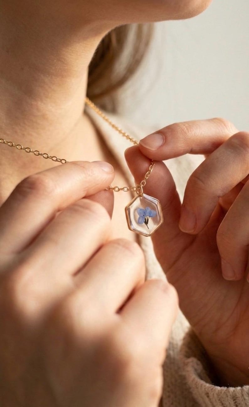 May include: A gold-colored necklace with a hexagonal pendant. The pendant encases a small, delicate blue flower in clear resin. The necklace is being held by a person.
