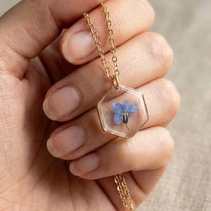 May include: A gold-colored necklace featuring a hexagonal pendant with a small blue flower encased in clear resin. The delicate chain and pendant are held in a hand, showcasing the jewelry's intricate design and floral element.