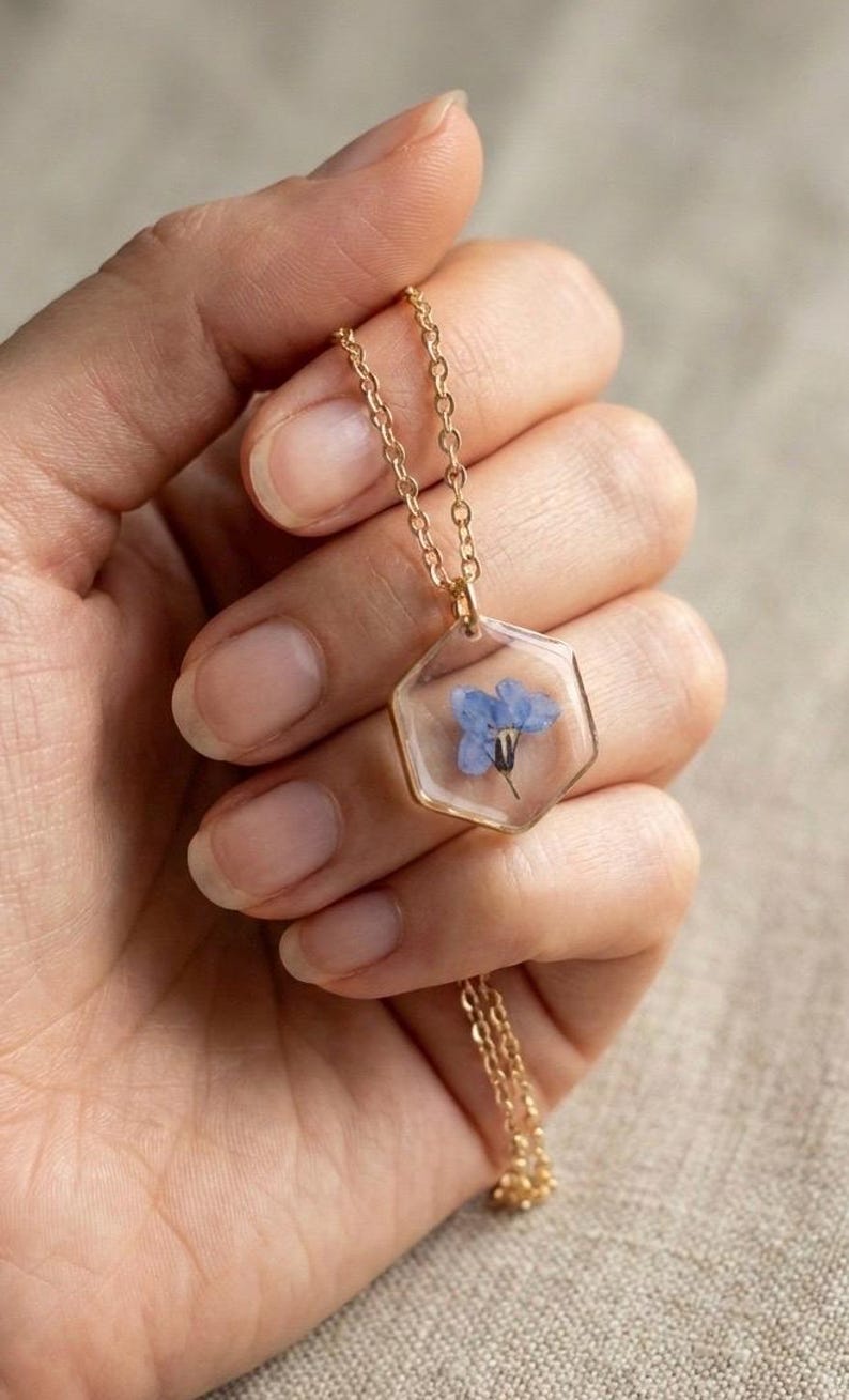 May include: A gold-colored necklace featuring a hexagonal pendant with a small blue flower encased in clear resin. The delicate chain and pendant are held in a hand, showcasing the jewelry's intricate design and floral element.