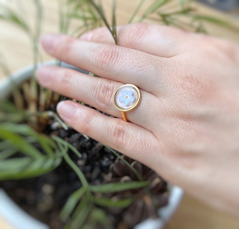 May include: A gold ring with a clear resin setting featuring a delicate blue flower.