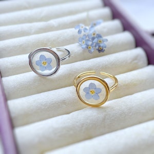 May include: Two silver and gold rings with a delicate blue flower pressed inside a clear glass dome. The rings are displayed in a white velvet ring box.