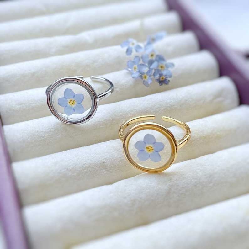 May include: Two silver and gold rings with a delicate blue flower pressed inside a clear glass dome. The rings are displayed in a white velvet ring box.
