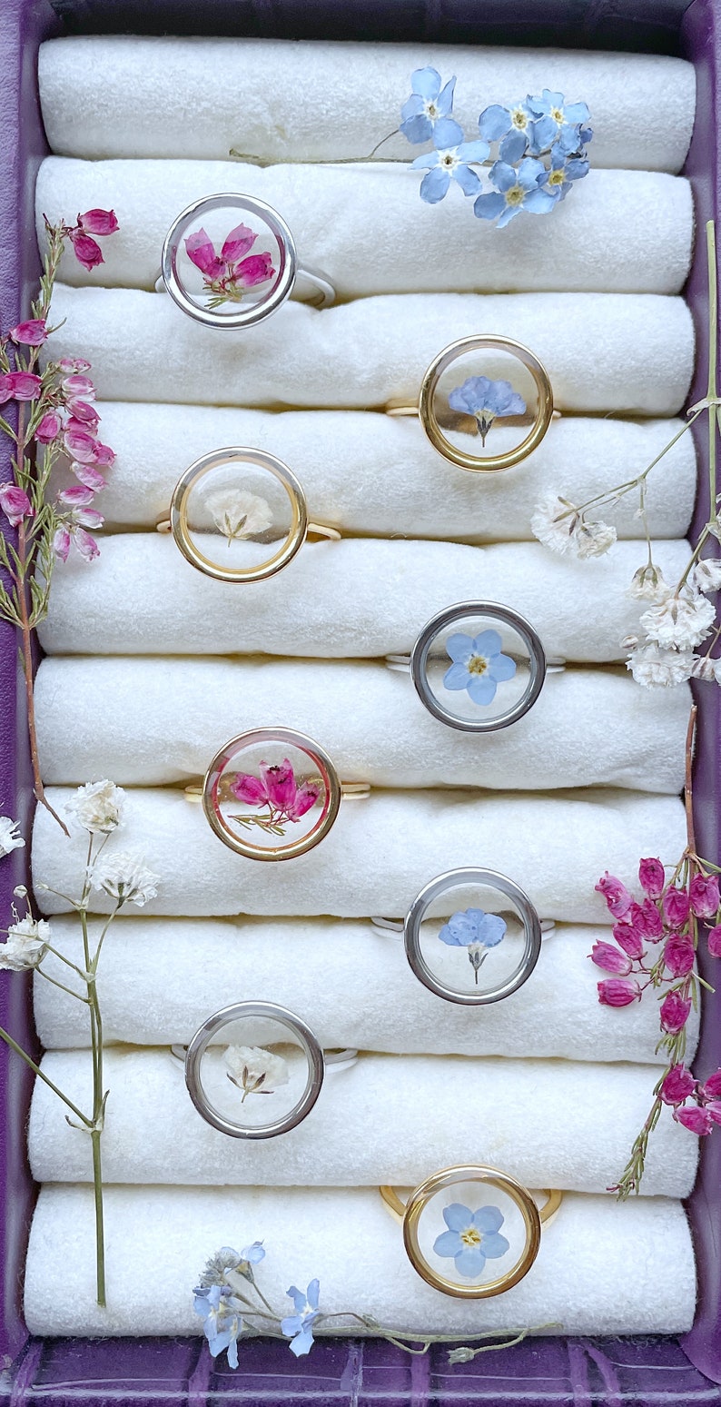 May include: A collection of eight rings with different pressed flowers inside. The rings are displayed in a purple velvet box with white fabric lining. The flowers include pink, white, and blue.