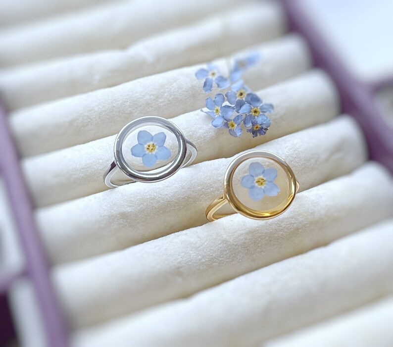 May include: Two rings with forget-me-not flowers encased in clear resin. One ring is silver and the other is gold.