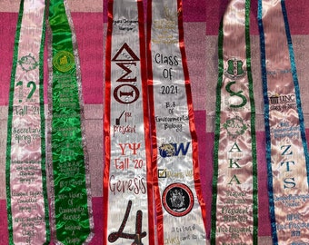 May include: Four graduation stoles with different colors and designs. The stoles are white with red, green, pink, and blue trim. Each stole has embroidered text and designs, including Greek letters, organization names, and graduation year.