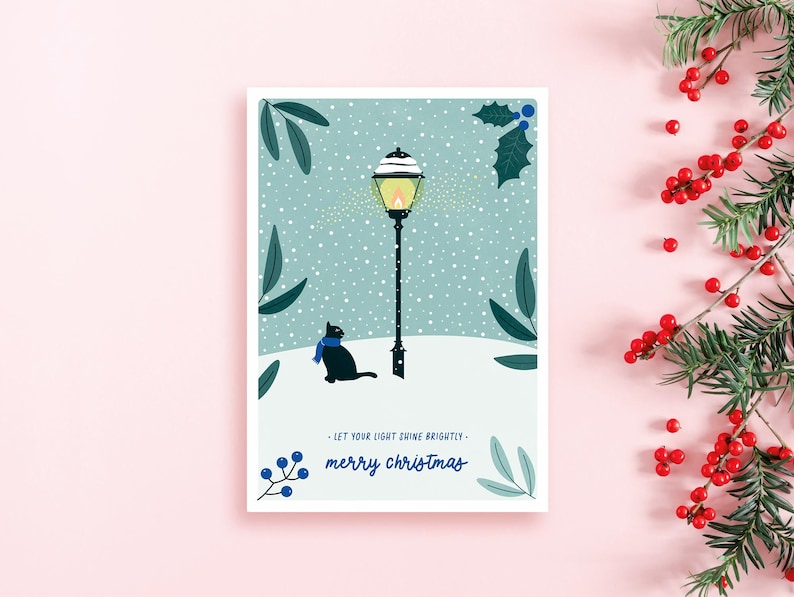 Auf der Weihnachtskarte aus nachhaltigen 100 % Recyclingpapier ist eine Katze abgebildet. Sie schaut in einer verschneiten Winterlandschaft zu einer leuchtenden Laterne. Die Farben: salbeigrün & blau. Let your light shine brightly. Merry Christmas