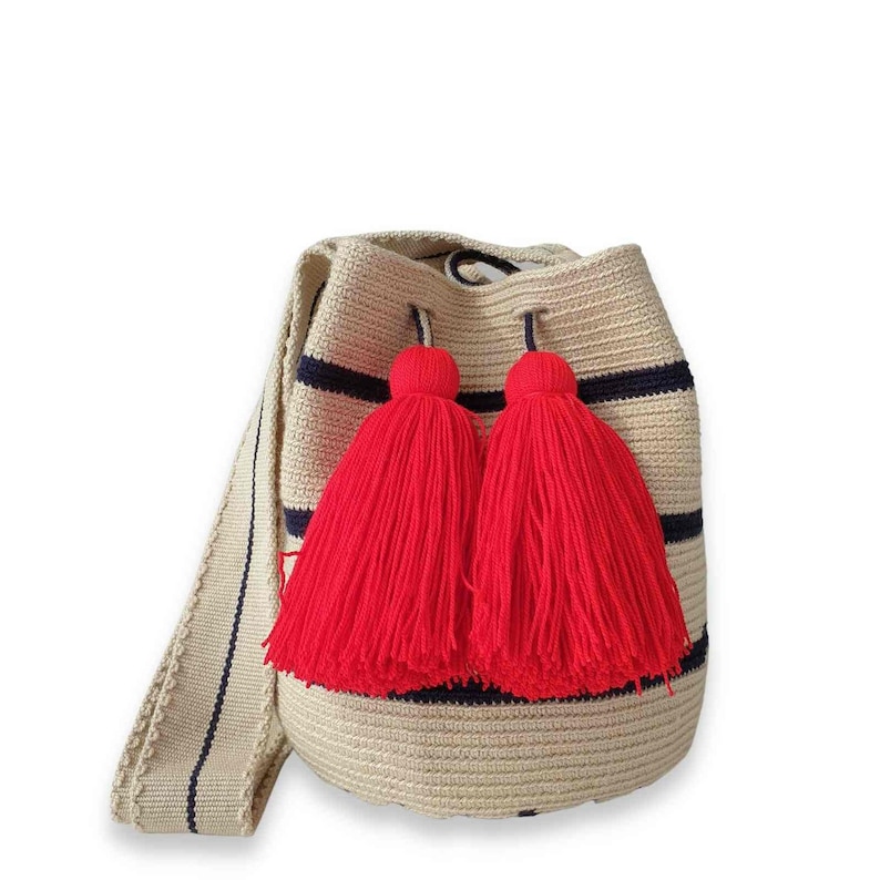 May include: A beige bucket bag with horizontal navy blue stripes, featuring two large, vibrant red yarn tassels hanging from the drawstring closure. The bag has a wide, matching beige shoulder strap with a thin navy blue center stripe.