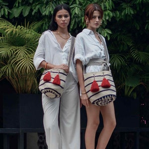May include: Two off-white bucket bags featuring horizontal navy blue stripes, a circular navy blue geometric pattern at the base, and two prominent red tassels hanging from the drawstring closure, each attached to a long, woven shoulder strap.