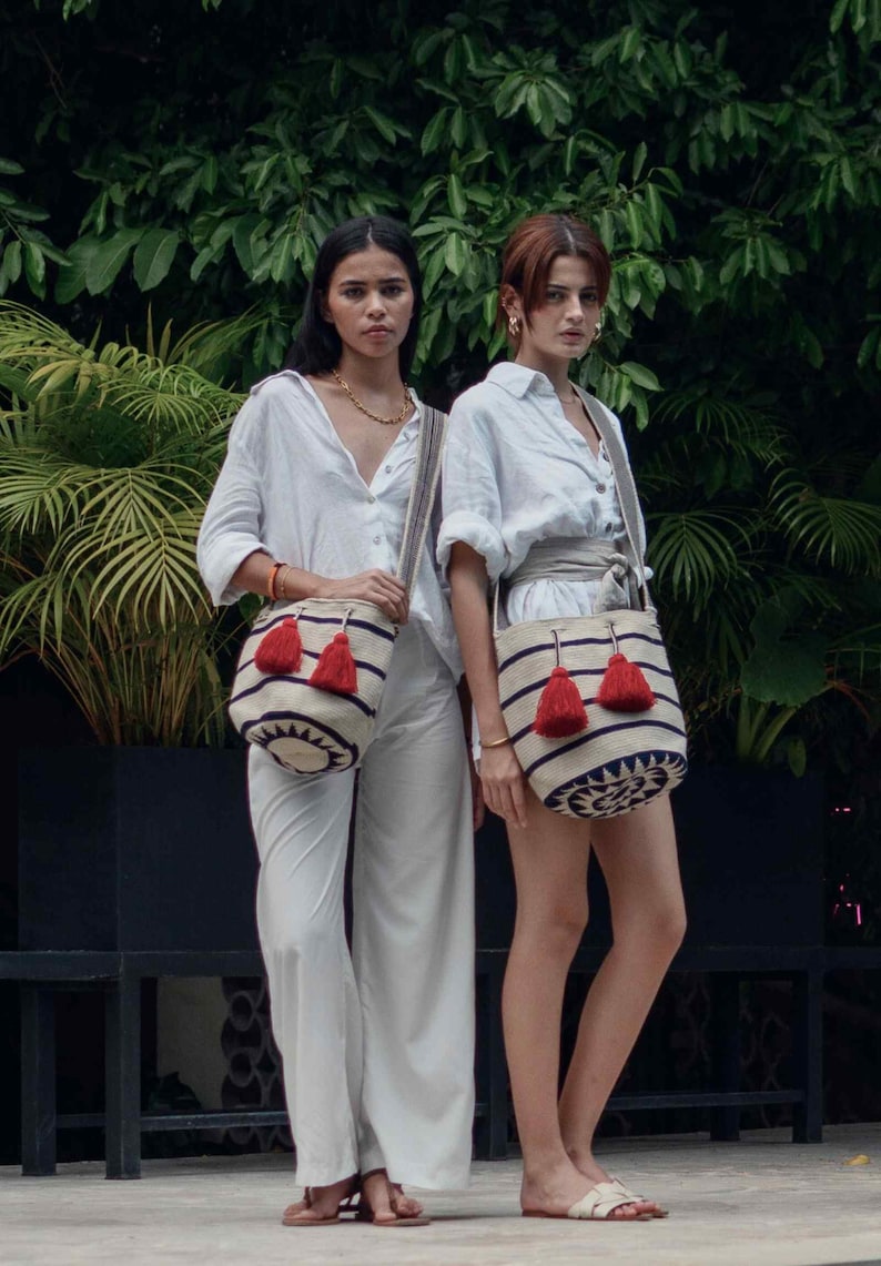 May include: Two off-white bucket bags featuring horizontal navy blue stripes, a circular navy blue geometric pattern at the base, and two prominent red tassels hanging from the drawstring closure, each attached to a long, woven shoulder strap.