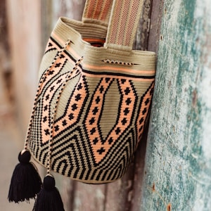 May include: A woven bucket bag featuring a geometric pattern in olive green, black, and peach. The bag has a wide shoulder strap, a drawstring closure with two long black tassels, and a structured cylindrical base with intricate thread work.