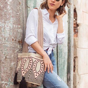 May include: A woven bucket bag featuring a geometric pattern in beige, black, and soft peach. The bag has a wide, striped shoulder strap and two black tassels hanging from the drawstring closure. The design includes repeating diamond shapes.