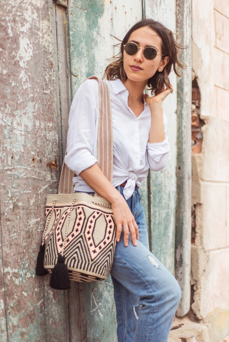 May include: A woven bucket bag featuring a geometric pattern in beige, black, and soft peach. The bag has a wide, striped shoulder strap and two black tassels hanging from the drawstring closure. The design includes repeating diamond shapes.