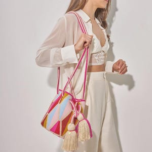 Op de afbeelding: Een kleurrijke geweven emmertas met een geometrisch patroon in roze, rood, geel en blauw. De tas heeft een roze band en twee grote beige kwasten. De tas wordt vastgehouden door een persoon die een wit overhemd en een broek draagt.
