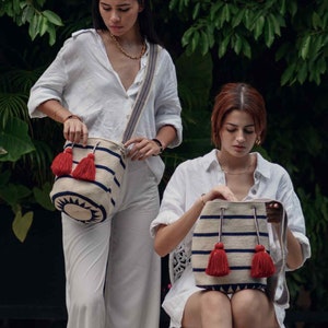 May include: A cylindrical woven shoulder bag featuring a cream-colored base with horizontal navy blue stripes, a dark navy blue geometric sunburst pattern at the base, and two prominent red tassels attached to the drawstring closure.