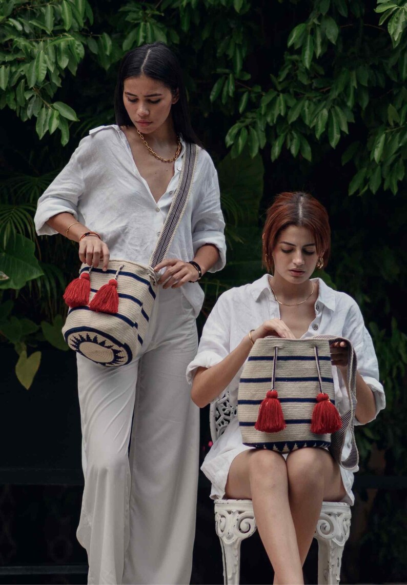 May include: A cylindrical woven shoulder bag featuring a cream-colored base with horizontal navy blue stripes, a dark navy blue geometric sunburst pattern at the base, and two prominent red tassels attached to the drawstring closure.