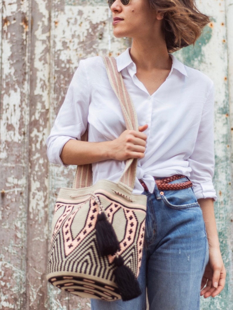 May include: A bucket bag featuring a woven geometric pattern in beige, black, and soft pink. The bag has a wide shoulder strap, a drawstring closure, and two black tassels hanging from the side. The design includes intricate diamond shapes.