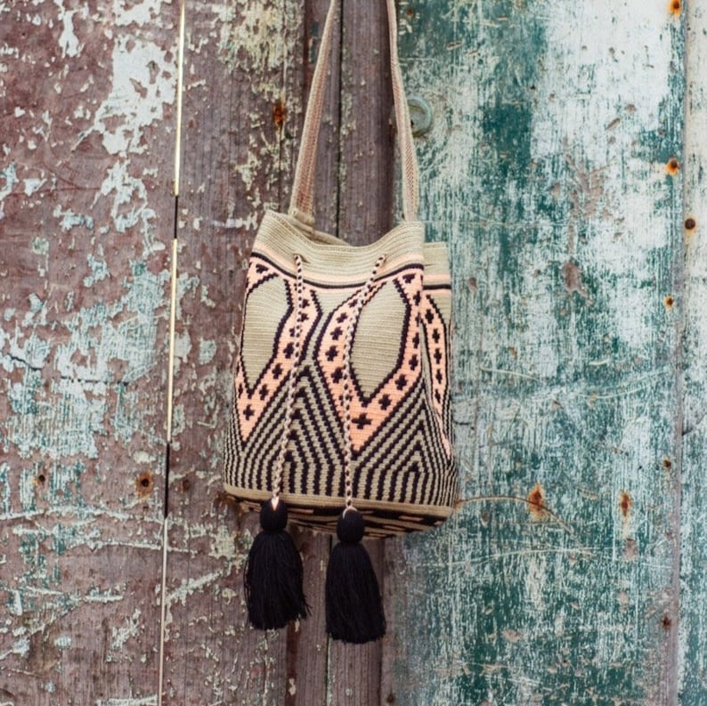 May include: A bucket bag featuring a woven geometric pattern in beige, black, and soft peach. The bag includes a long shoulder strap, a drawstring closure with braided cords, and two large black tassels hanging from the front of the bag.