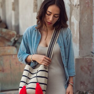 May include: A woven bucket bag featuring horizontal black stripes on a cream background, a wide shoulder strap with a black and white pattern, and two long drawstrings ending in large, vibrant red tassels hanging from the front of the bag.