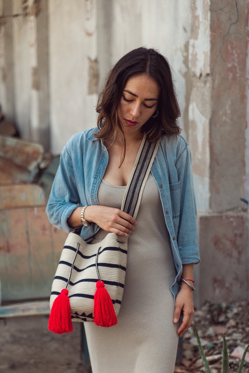May include: A woven bucket bag featuring horizontal black stripes on a cream background, a wide shoulder strap with a black and white pattern, and two long drawstrings ending in large, vibrant red tassels hanging from the front of the bag.