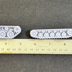 Puede incluir: Cuatro conjuntos de orugas de tanque en miniatura de color gris claro, de diversas longitudes y dise&ntilde;os, alineados sobre una regla amarilla de 30 cm. Los componentes muestran ruedas, pi&ntilde;ones y patrones de oruga detallados en material r&iacute;gido.