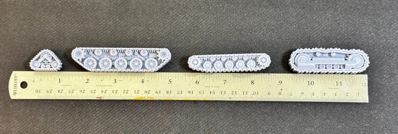 Puede incluir: Cuatro conjuntos de orugas de tanque en miniatura de color gris claro, de diversas longitudes y dise&ntilde;os, alineados sobre una regla amarilla de 30 cm. Los componentes muestran ruedas, pi&ntilde;ones y patrones de oruga detallados en material r&iacute;gido.