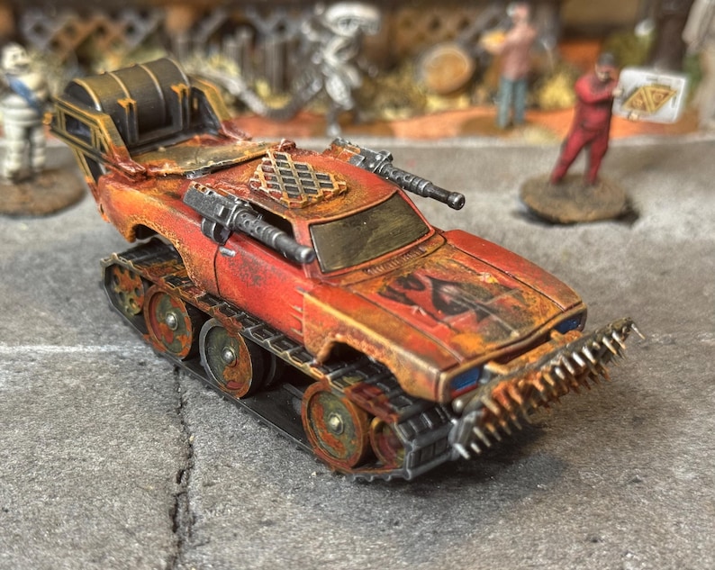Puede incluir: Un veh&iacute;culo en miniatura postapocal&iacute;ptico con pintura desgastada en rojo y naranja, orugas de tanque, parachoques delantero con p&uacute;as, dos ca&ntilde;ones laterales, una rejilla en el techo y un gran tambor de carga trasero, con detalles de &oacute;xido.