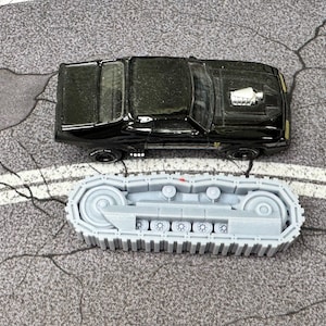 Puede incluir: Un coche de juguete tipo muscle car negro con un detalle de motor plateado en el cap&oacute;, colocado sobre un componente de oruga de tanque de pl&aacute;stico gris claro con una serie de ruedas peque&ntilde;as y un dise&ntilde;o de banda continua para veh&iacute;culos.
