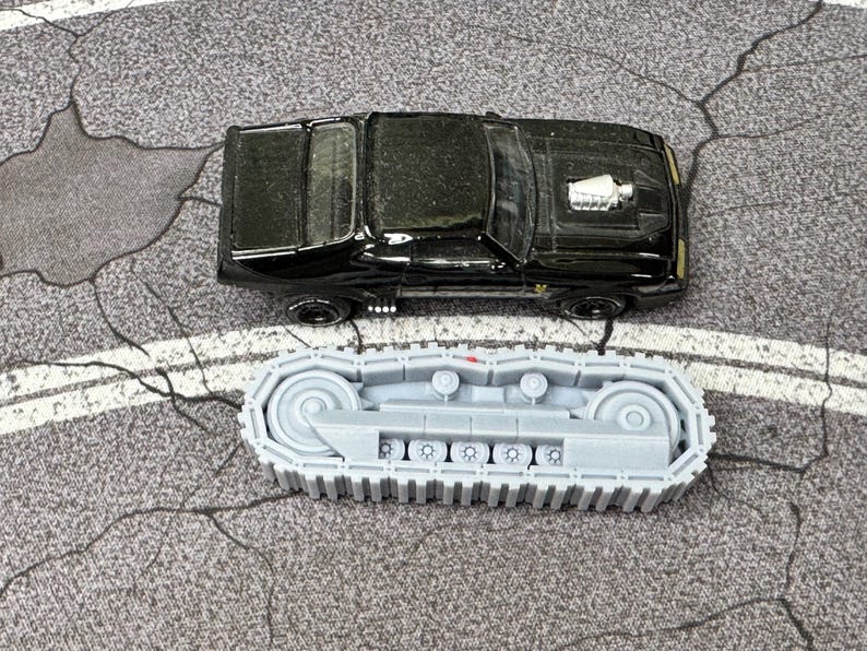 Puede incluir: Un coche de juguete tipo muscle car negro con un detalle de motor plateado en el cap&oacute;, colocado sobre un componente de oruga de tanque de pl&aacute;stico gris claro con una serie de ruedas peque&ntilde;as y un dise&ntilde;o de banda continua para veh&iacute;culos.
