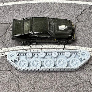 Puede incluir: Un coche de juguete negro con un detalle de motor plateado en el cap&oacute;, colocado sobre un conjunto de orugas de tanque de pl&aacute;stico gris claro que presenta m&uacute;ltiples ruedas circulares y un dise&ntilde;o de oruga continuo sobre una superficie gris.
