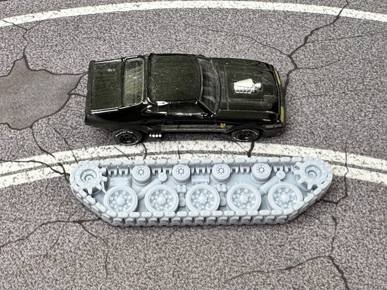 Puede incluir: Un coche de juguete negro con un detalle de motor plateado en el cap&oacute;, colocado sobre un conjunto de orugas de tanque de pl&aacute;stico gris claro que presenta m&uacute;ltiples ruedas circulares y un dise&ntilde;o de oruga continuo sobre una superficie gris.