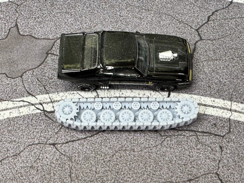 Puede incluir: Un coche de juguete negro tipo muscle car con un detalle de motor plateado en el cap&oacute;, situado sobre un componente de oruga de tanque gris claro alargado con m&uacute;ltiples ruedas circulares peque&ntilde;as y un dise&ntilde;o de cadena para modelos.